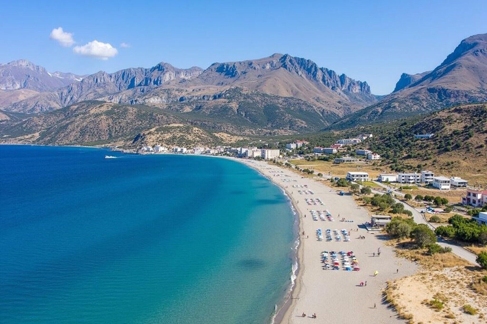 Una playa costera con mar azul cristalino, arena dorada y montañas al fondo.