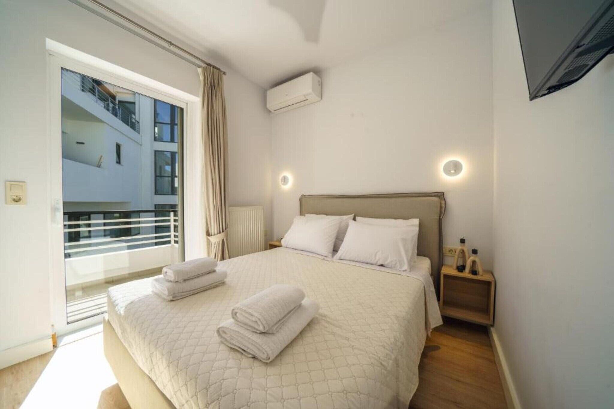 Un dormitorio con cama, almohadas, mesita de noche, aire acondicionado instalado en la pared y ventana con cortinas.