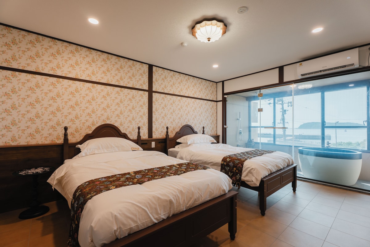Habitación de hotel con dos camas, papel tapiz floral, un ventanal con vistas y un baño moderno.
