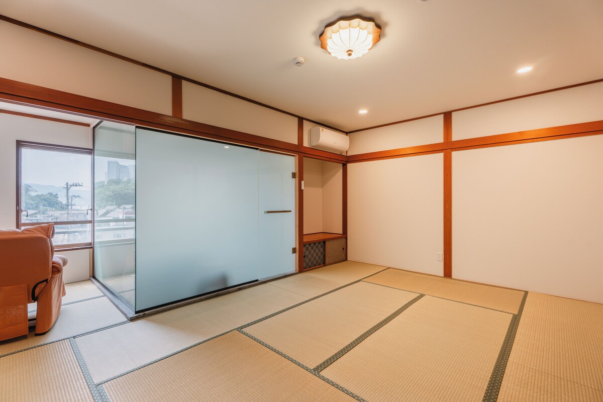 Una habitación con suelo de tatami, puerta corredera de vidrio, un sillón acolchado y una lámpara en el techo.