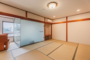 Una habitación con suelo de tatami, puerta corredera de vidrio, un sillón acolchado y una lámpara en el techo.