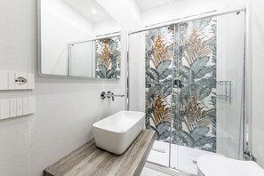 Ein modernes Badezimmer mit einer freistehenden Badewanne, einer Duschkabine mit tropischen Blattmustern und einem großen Spiegel über dem Waschbecken.