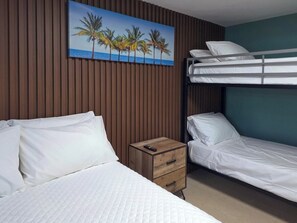 A hotel room with a bunk bed, a single bed, a wooden nightstand with a remote control, and a painting of palm trees on the wall. - Bahía Bonita | Cozy Cabo Rojo Retreat (Cabo Rojo)