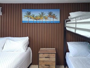 Ein Hotelzimmer mit zwei Betten, einem Holfnachtkästchen und einem Palmenbild.