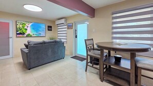 Una sala de estar moderna con un sofá, una mesa de comedor con sillas, un televisor montado en la pared y una puerta que da al exterior.