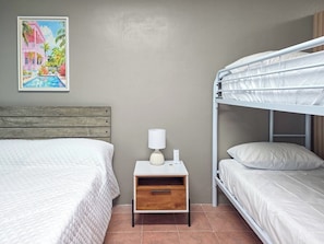 A bedroom with a bunk bed, a single bed, a nightstand with a lamp, and a painting on the wall. - Brisa Del Caribe | Top Floor Studio in Cabo Rojo (Cabo Rojo)