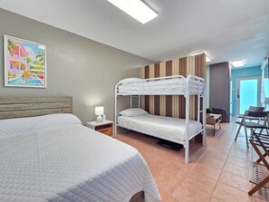 A hotel room with a bunk bed, a single bed, a desk with a chair, a lamp, and a painting on the wall. - Brisa Del Caribe | Top Floor Studio in Cabo Rojo (Cabo Rojo)
