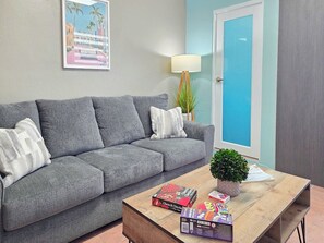 A living room with a grey sofa, a wooden coffee table with board games, a small potted plant, and a lamp. - Brisa Del Caribe | Top Floor Studio in Cabo Rojo (Cabo Rojo)