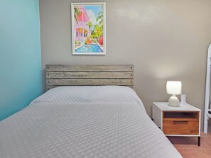 Una cama con cubrecama blanco acolchado, un cabecero de madera, una mesita de noche con una lámpara blanca y un cuadro enmarcado de una casa rosa con piscina.
