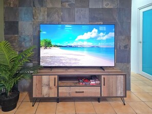 Ein Flachbildfernseher auf einem hölzernen TV-Schrank, auf dem eine Strandansicht zu sehen ist, eine Topfpflanze links daneben und ein gefliester Boden.