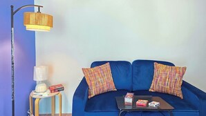 A blue sofa with two patterned pillows, a small table with books and a box, a floor lamp, and a wall-mounted lamp. - Palmera Azul | Cozy Private Apartment in Cabo Rojo (Cabo Rojo)