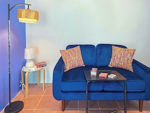 A blue sofa with two patterned pillows, a small table with books and a box, a floor lamp, and a wall-mounted lamp. - Palmera Azul | Cozy Private Apartment in Cabo Rojo (Cabo Rojo)