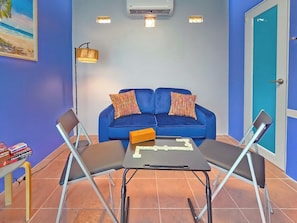 A room with a blue sofa, two chairs, a table with dominoes, a lamp, and a door. - Palmera Azul | Cozy Private Apartment in Cabo Rojo (Cabo Rojo)