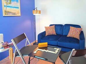 Een kamer met een blauwe bank, twee gestreepte kussens, een klein tafeltje met een spel, een staande lamp en een lijst met een afbeelding aan de muur.