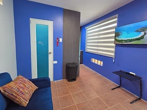 Ein Zimmer mit blauer Wand, einer Tür, einem kleinen Mülleimer, einem blauen Sofa mit farbigem Kissen, einem schwarzen Tisch und einem Fenster mit Jalousien.