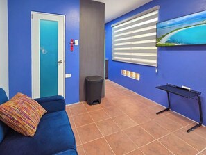 A room with a blue wall, a door, a trash bin, a blue sofa with a colorful cushion, a small table, and a window with blinds. - Villa Andrea | Cabo Rojo Private Estate (Cabo Rojo)