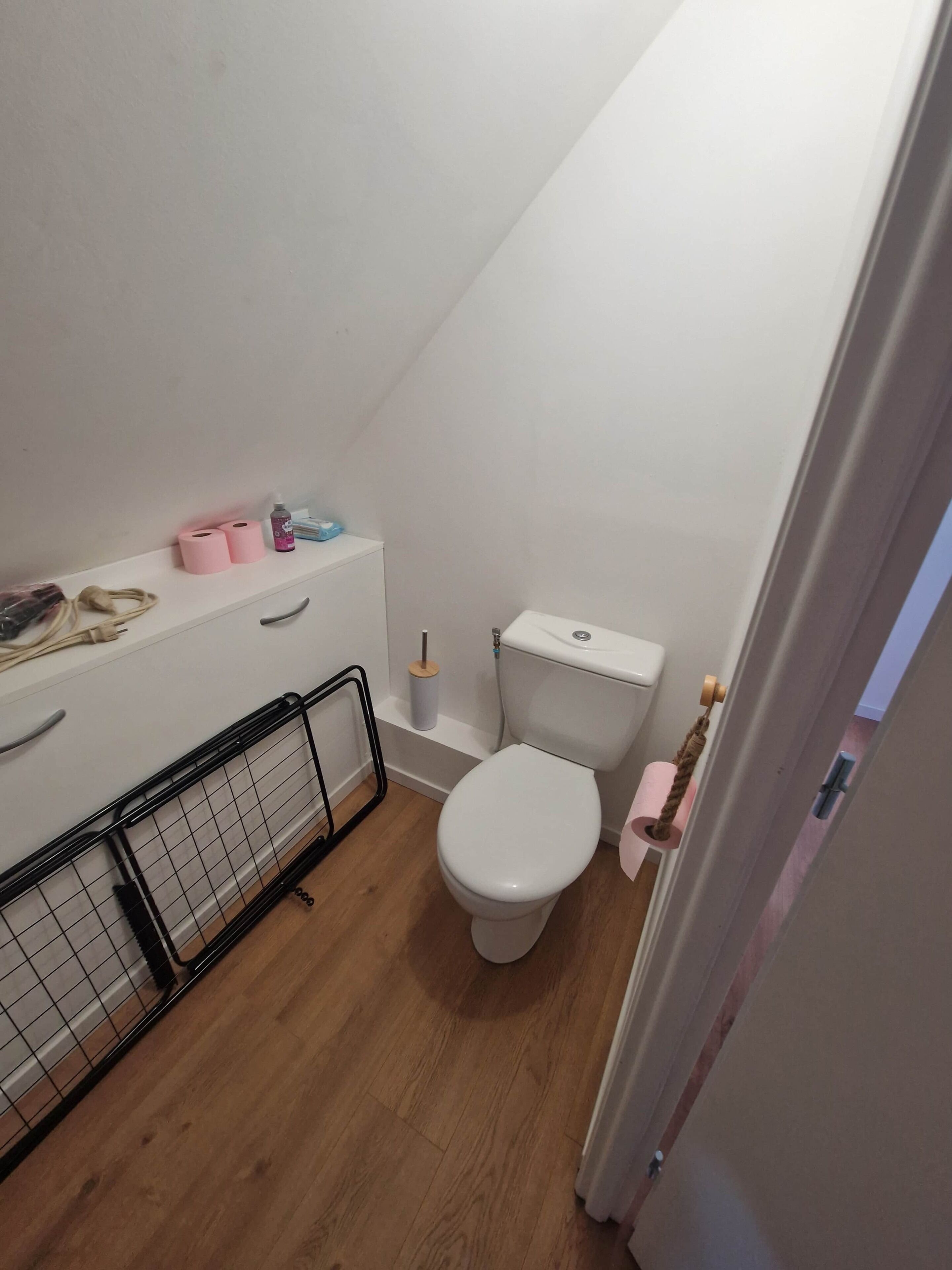 A small, compact bathroom with a toilet, a shelf with toiletries, and a wooden floor.