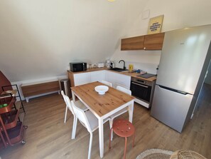 Uma cozinha compacta com mesa de madeira, cadeiras brancas, micro-ondas, máquina de café e geladeira.