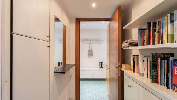 Un pasillo con una estantería, una puerta que da al baño y suelo de baldosas.