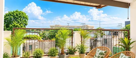Un balcon agrémenté de fauteuils en osier, de plantes en pot et offrant une vue sur les immeubles et les arbres.