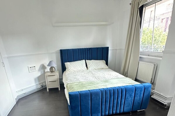 A bedroom with a bed, a nightstand, a lamp, and a window. - Cozy Apartment in Central London by Tower Bridge (London)