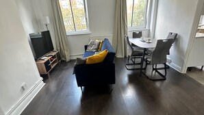 Ein modernes Wohnzimmer mit dunklem Holzboden, einem blauen Sofa mit gelben Kissen, einem TV-Schrank und einem Essbereich mit Tisch und Stühlen.