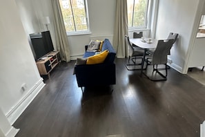 Ein modernes Wohnzimmer mit dunklem Holzboden, einem blauen Sofa mit gelben Kissen, einem TV-Schrank und einem Essbereich mit Tisch und Stühlen.