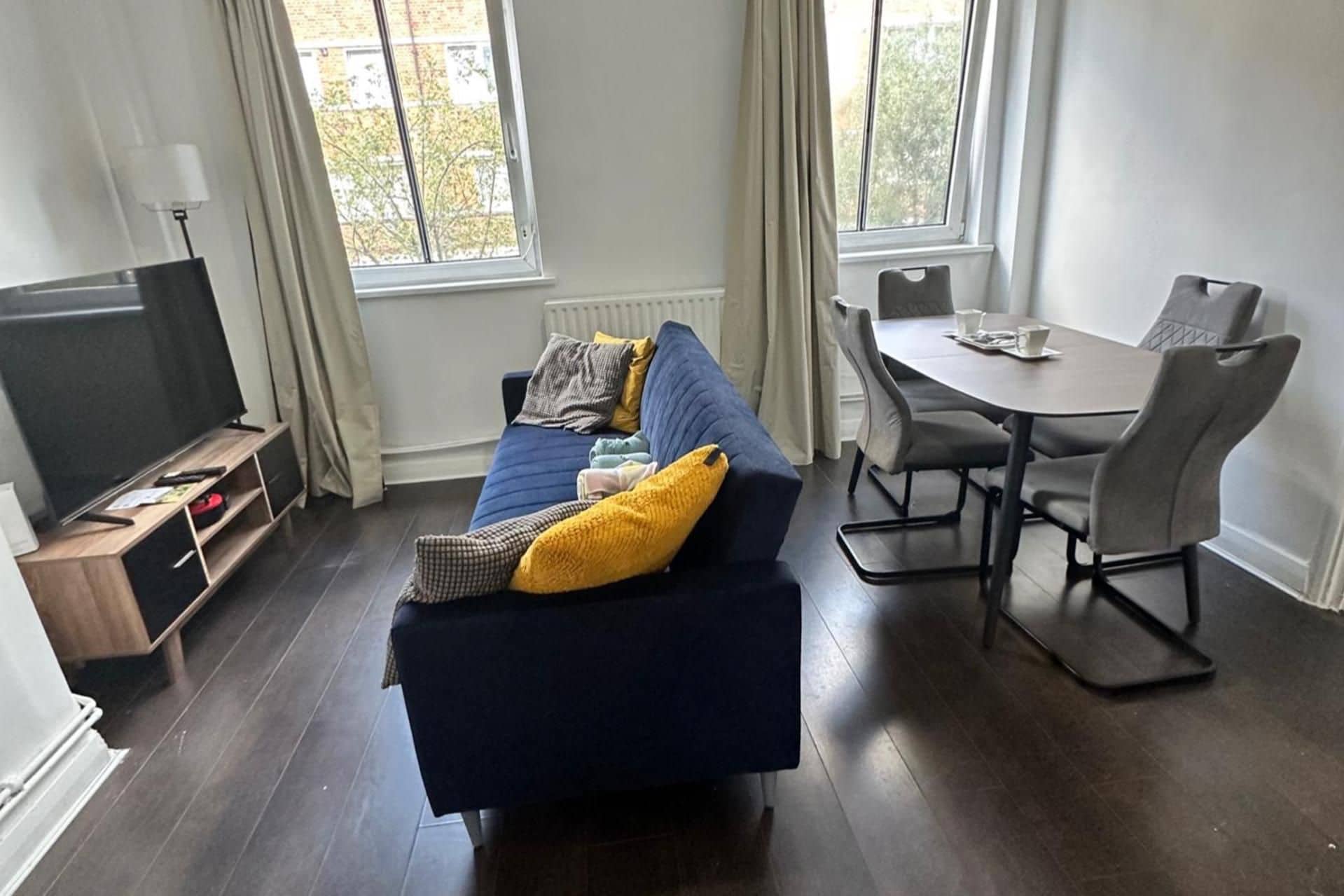 Ein modernes Wohnzimmer mit einem blauen Sofa, einem Esstisch mit Stühlen, einem Flachbildfernseher und einem Fenster mit Vorhängen.