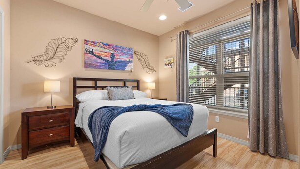 Quarto com cama, mesas de cabeceira, ventoinha no teto, janela com persianas e quadros nas paredes.