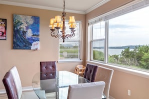 A dining area with a glass table, cushioned chairs, a chandelier, a painting, and a view of the outdoors. - The Laughing Pointe Lakeview (Hollister)