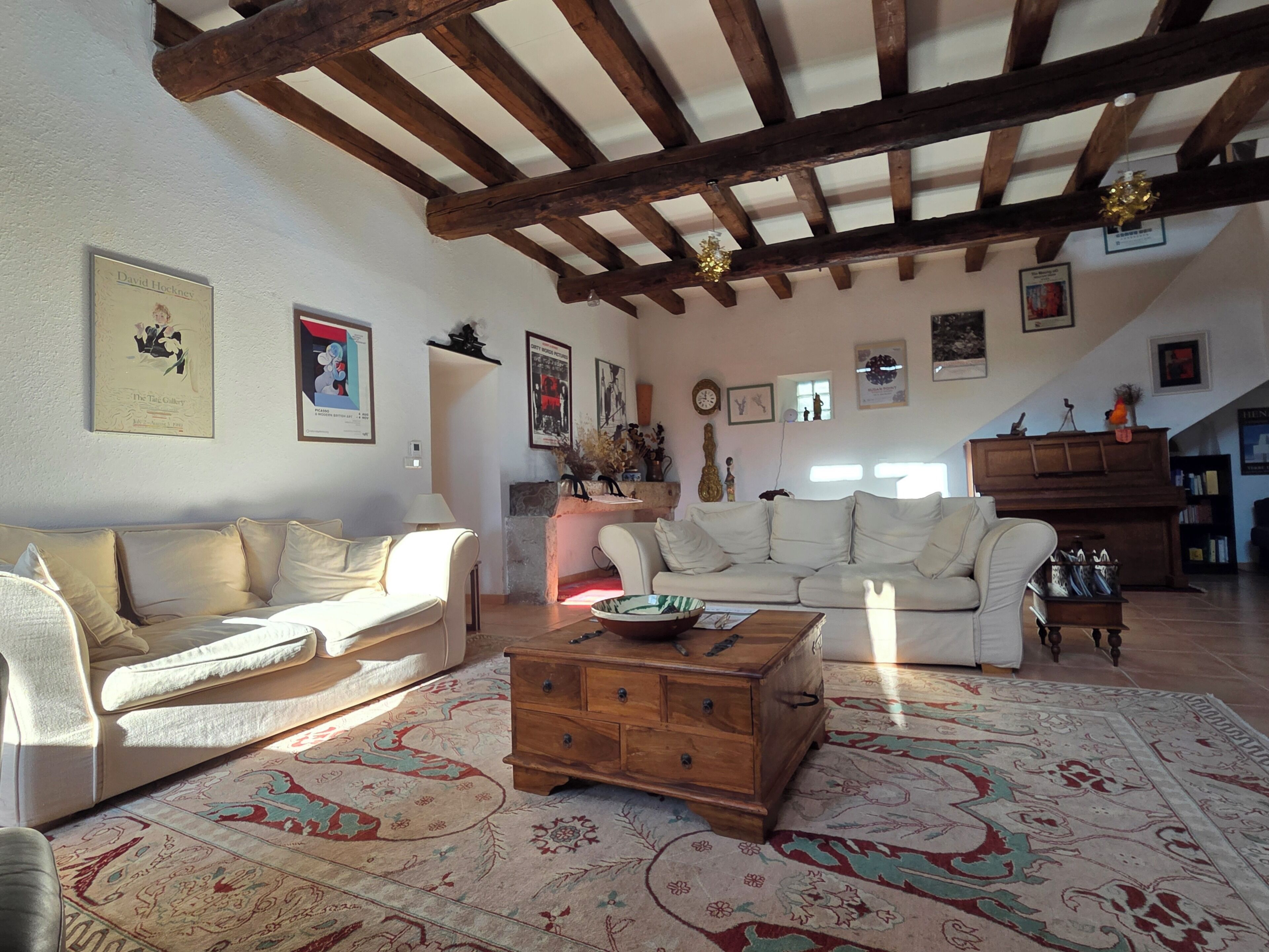 A spacious living room with a high ceiling and exposed wooden beams, furnished with a beige sofa, a white armchair, and a wooden coffee table.