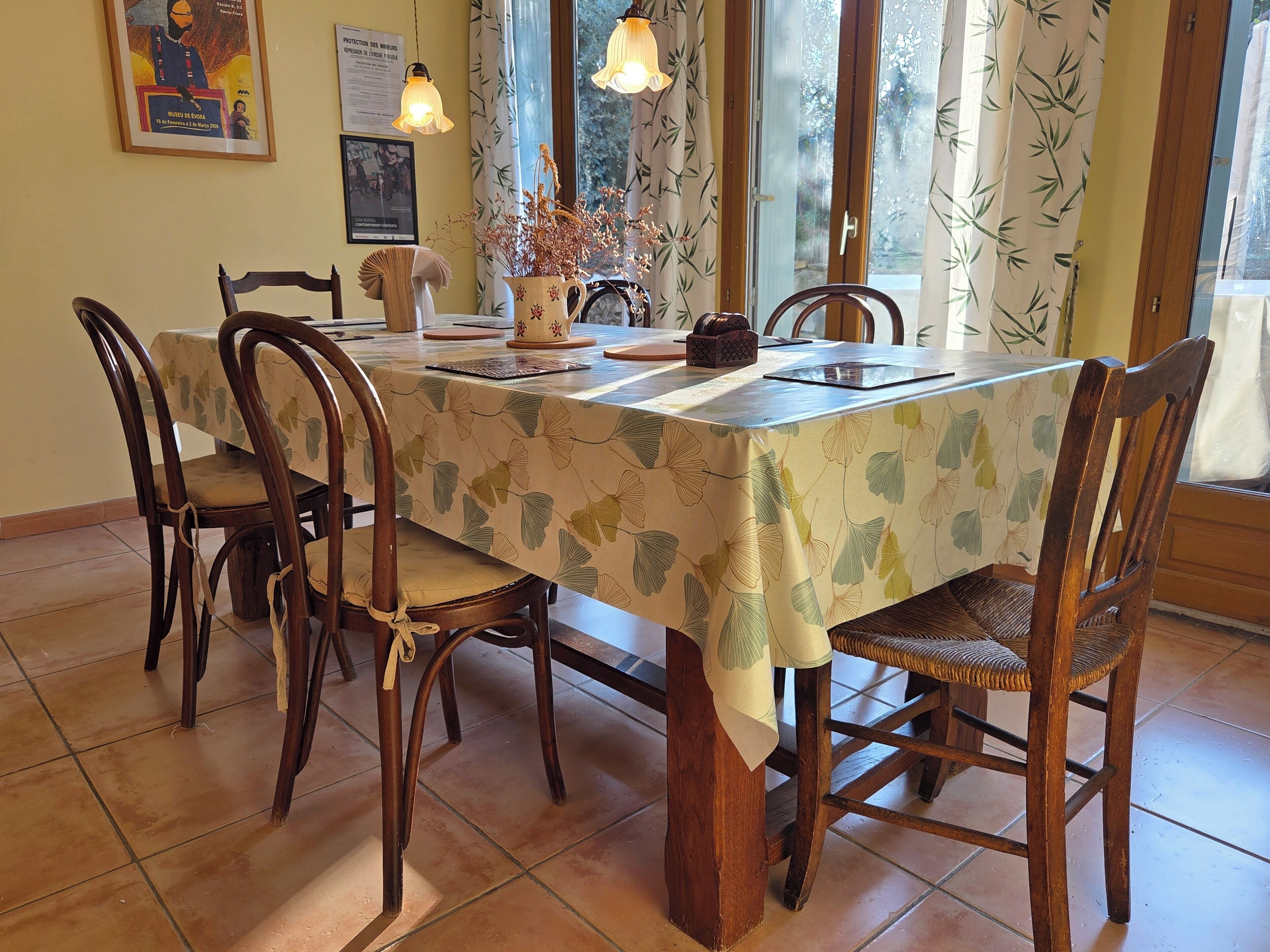 A dining area with a table covered in a leaf-patterned tablecloth, surrounded by wooden chairs with cushioned seats.