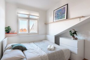 A bedroom with a bed, a radiator, a window, a shelf with plants, and a framed picture on the wall. - Bright Duplex Loft near Wenceslas Square (Prague 2)