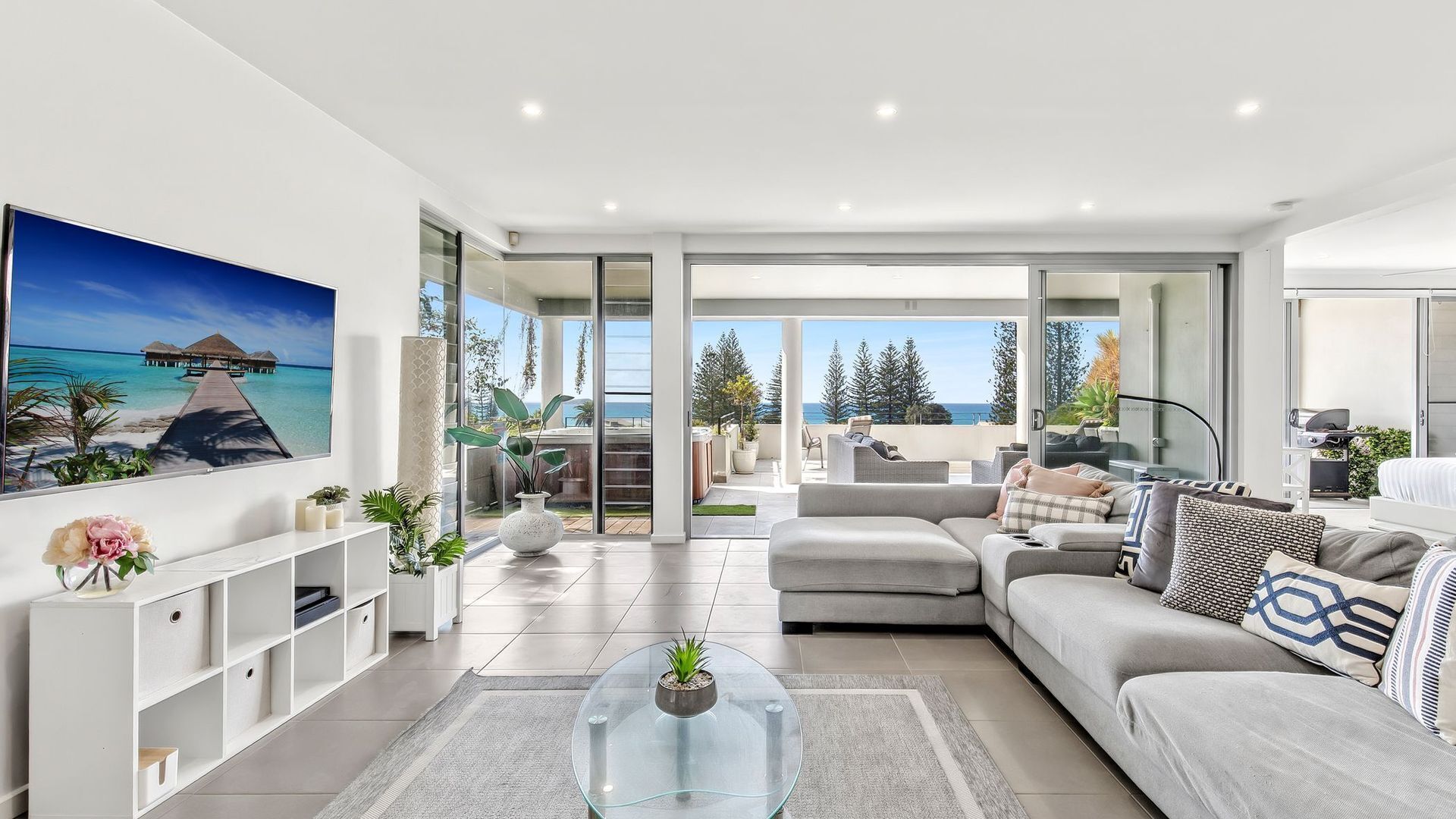 A modern living room with a large sofa, a glass coffee table, and a view of the ocean through large sliding doors.