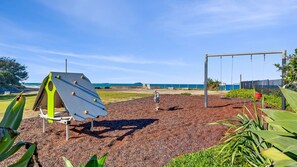 Ein Spielplatz mit Klettergerüst und Schaukeln, umgeben von einer Grünfläche mit Pflanzen.