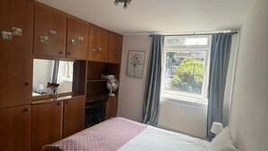 Ein Schlafzimmer mit einem Bett, einem Holzschrank, einem Fenster mit Vorhängen und einem Wandventilator.