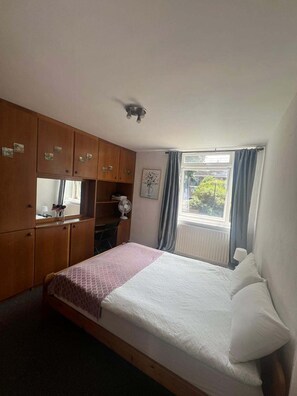 Ein Schlafzimmer mit einem Bett, einem Holzschrank, einem Fenster mit Vorhängen und einem Wandventilator.