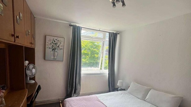 Ein Schlafzimmer mit Bett, Schreibtisch, Kleiderschrank, einem Fenster mit Vorhängen und einem Deckenventilator.