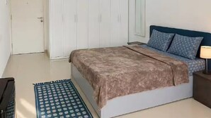 Un dormitorio con una cama, un armario, una mesita de noche con lámpara y una alfombra estampada.