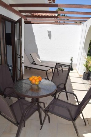 Un patio al aire libre con una mesa redonda de vidrio y cuatro sillas, un frutero y una pared blanca al fondo.