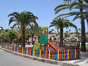 Un parque infantil con tobogán y estructuras para escalar, rodeado de una valla colorida y palmeras.