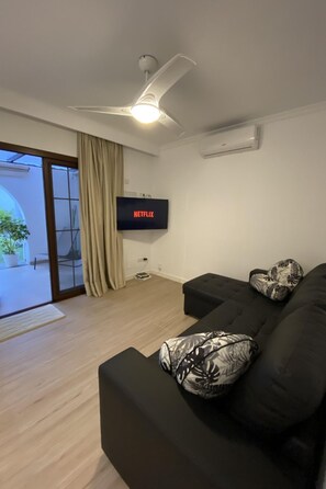 A modern living room with a black sofa, a ceiling fan, a flat-screen TV mounted on the wall, and a sliding glass door leading to an outdoor area. - für 4 Personen ca. 40 m² in Maspalomas, Gra by Interhome (Maspalomas)