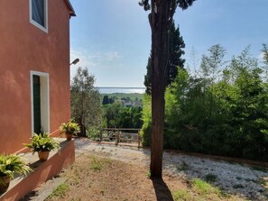 A house with a red exterior, a balcony with potted plants, a wooden fence, and a view of a green landscape and a body of water in the distance. - Collina Lagomare by Interhome (Corsanico)