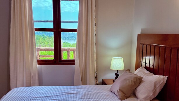A hotel room with a bed, a nightstand with a lamp, and a window with curtains. - Ewafe Safari Lodge (Paraa)