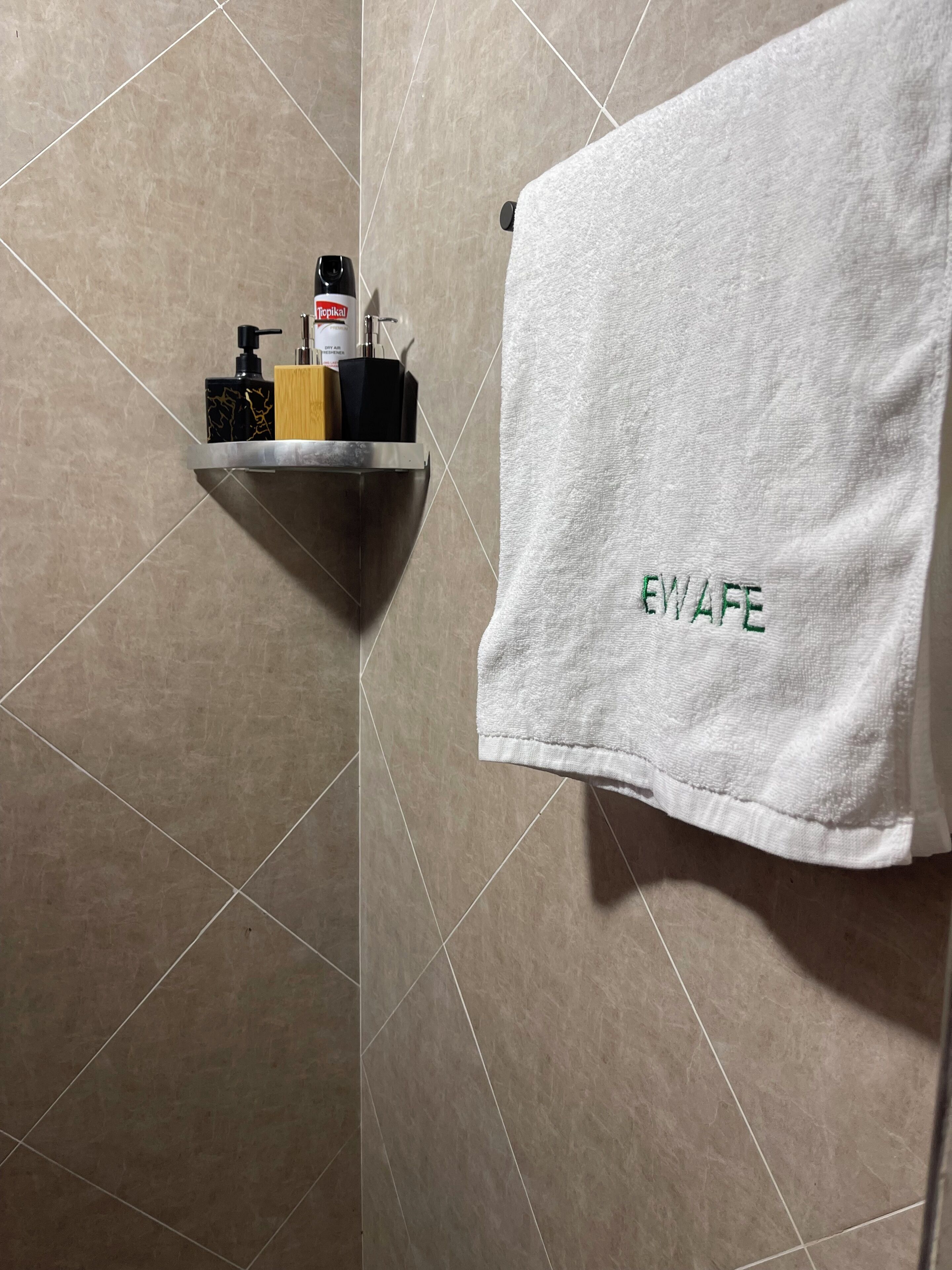 Een badkamer met een handdoekhouder met een witte handdoek waarop 'EWAFE' geborduurd staat, een plank met toiletartikelen en een betegelde wand.