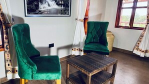 A room with two green upholstered chairs, a wicker table, a framed black and white photograph on the wall, and a window with curtains. - Ewafe Safari Lodge (Paraa)