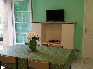 Ein Essbereich mit einem Tisch, der mit einer grünen Tischdecke bedeckt ist, einer Vase mit Blumen und einem Fernseher, der über einem Kamin an der Wand befestigt ist.