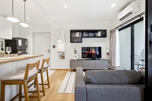 A modern living room with a grey sofa, wooden dining chairs, a kitchen area with a black refrigerator, and a mounted TV displaying 'NETFLIX'. - The Title Halo 1BR Apartment • Pools • 5 Min to Nai Yang Beach (Sakhu)