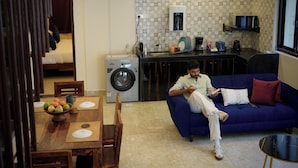 A living room with a blue sofa, a dining table with fruit, a washing machine, and a kitchen area with a coffee machine.