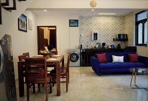 A living room with a blue sofa, a dining table with chairs, a washing machine, and a kitchen area with a microwave.